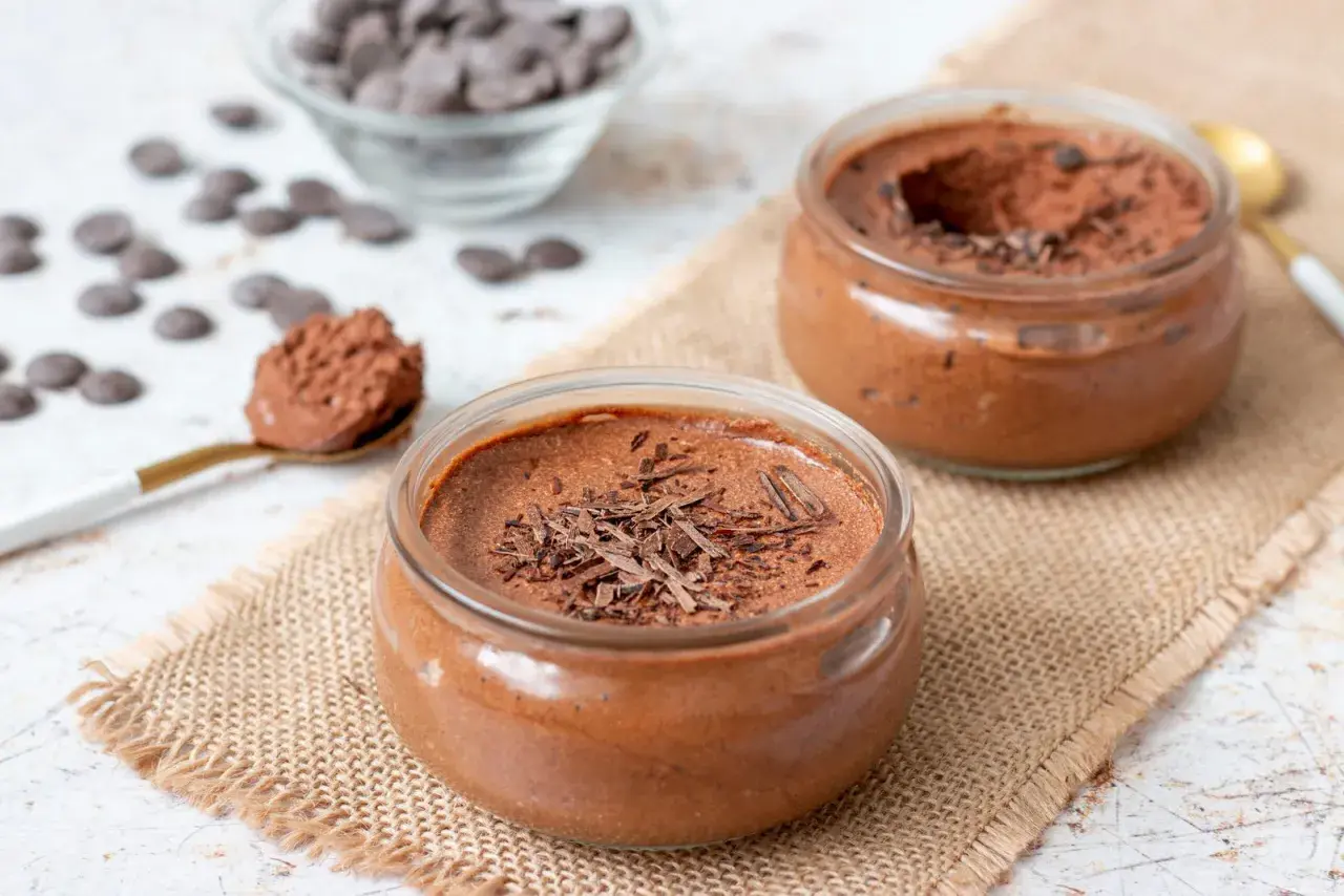 Deux pots de mousse au chocolat, garnie de copeaux de chocolat, sur une toile de jute. Une cuillère dorée est remplie de cette délicieuse mousse.