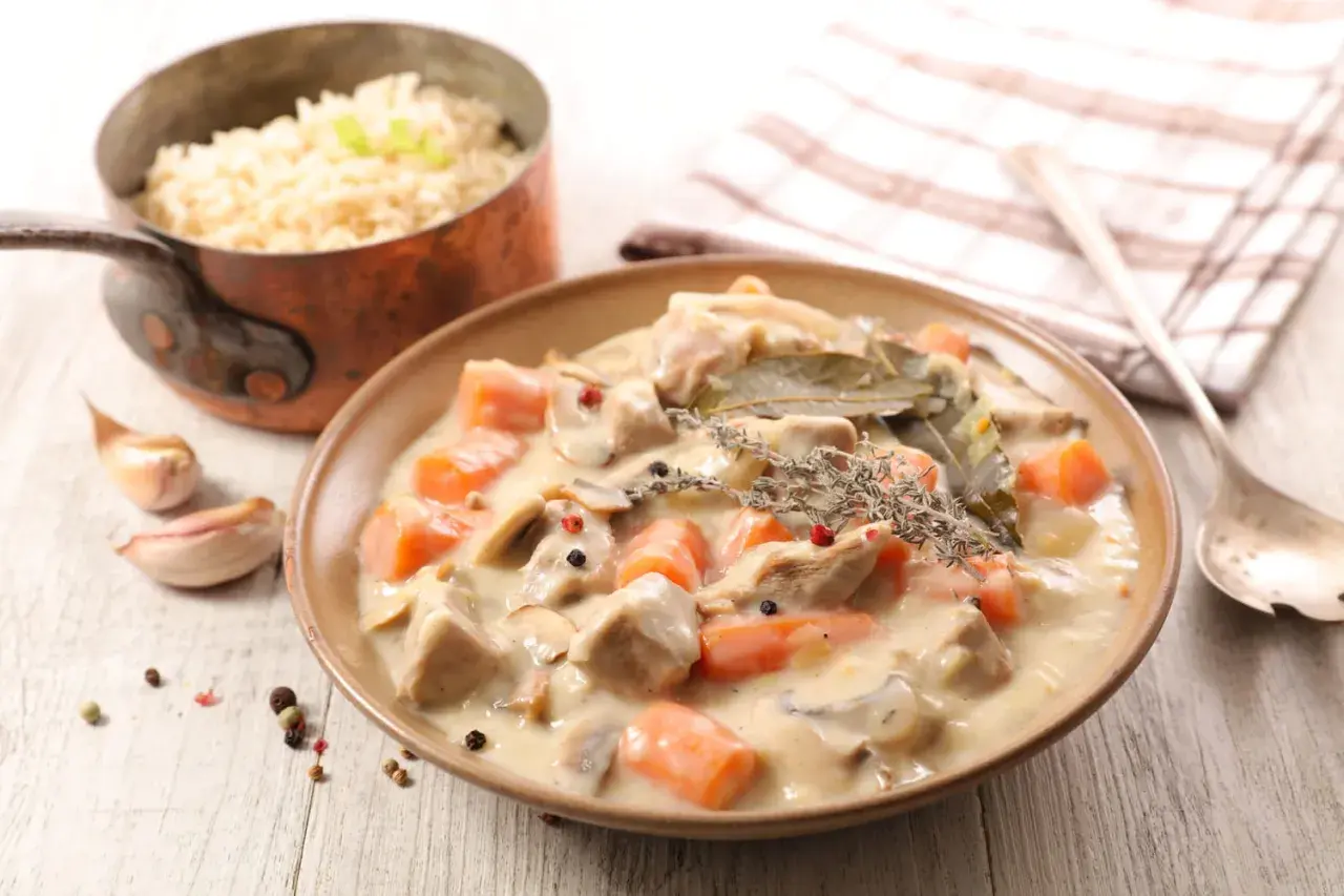 Un plat de blanquette de veau crémeuse avec des carottes et des champignons, garni de thym et de laurier. Du riz est servi à côté.