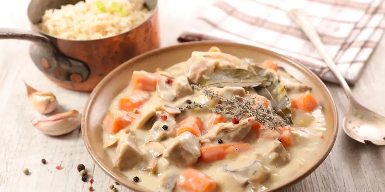 Un plat de blanquette de veau crémeuse avec des carottes et des champignons, garni de thym et de laurier. Du riz est servi à côté.