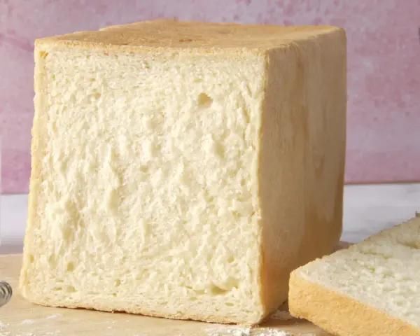 Un pain de mie moelleux, parfait pour une recette facile. Un pot de farine et un moule à pain sont à proximité.