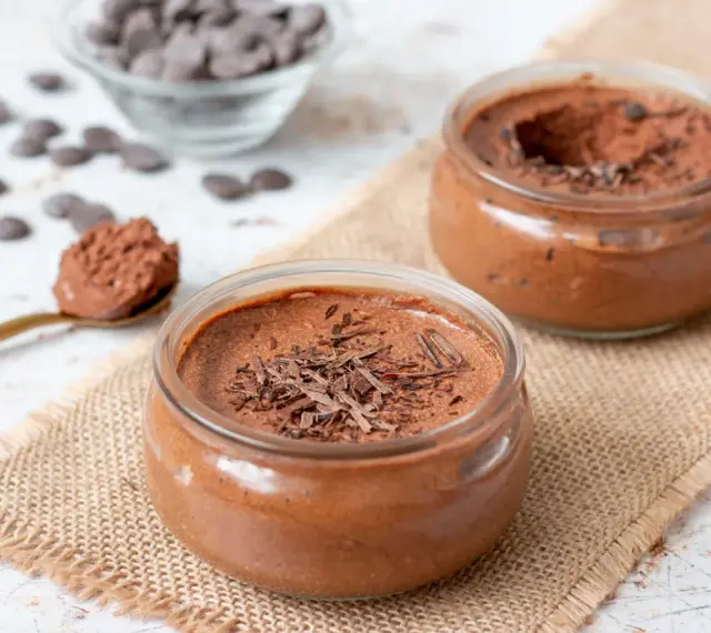 Deux pots de mousse au chocolat, garnie de copeaux de chocolat, sur une toile de jute. Une cuillère dorée est remplie de cette délicieuse mousse.