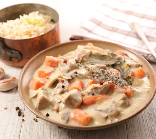 Un plat de blanquette de veau crémeuse avec des carottes et des champignons, garni de thym et de laurier. Du riz est servi à côté.