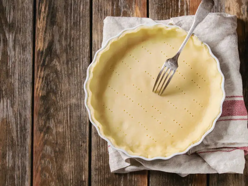 Pâte brisée sans beurre dans un plat à tarte, prête à être garnie. Une fourchette est plantée dans la pâte.