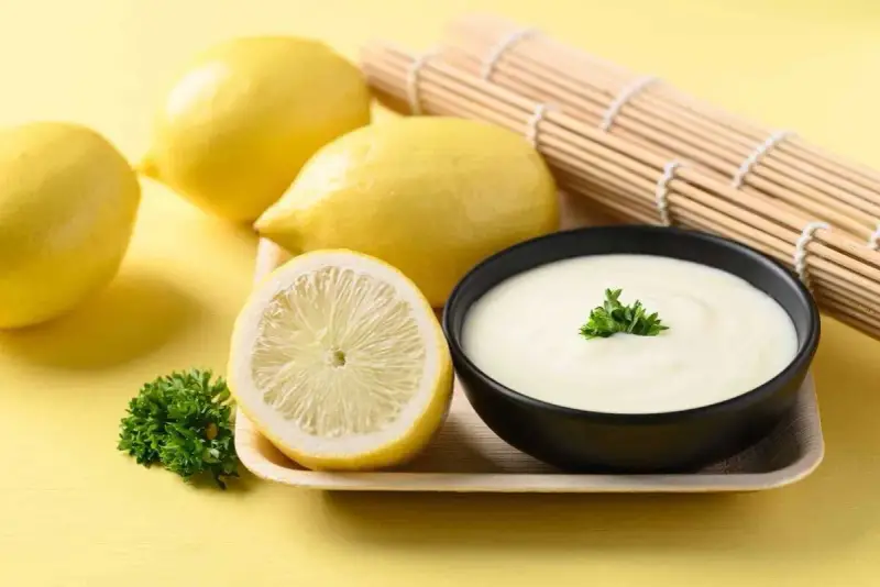 Un bol de sauce beurre citron crémeuse, garni de persil, avec des citrons frais et un rouleau de bambou en arrière-plan.