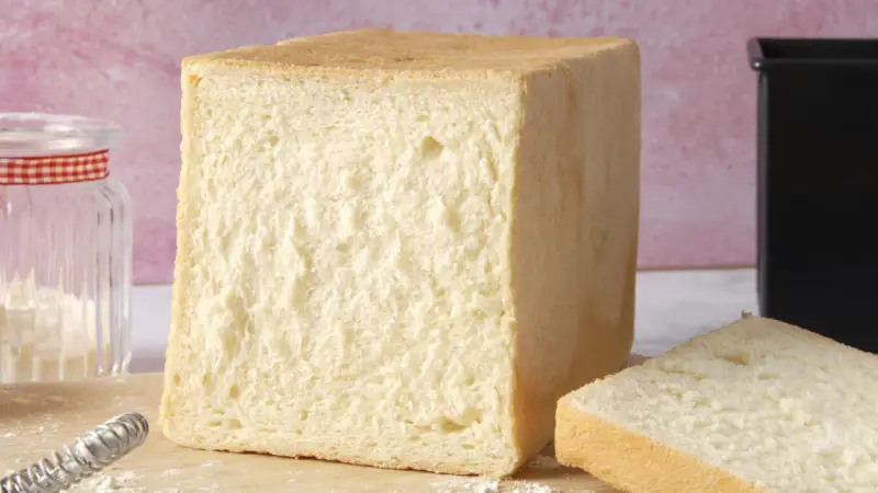 Un pain de mie moelleux, parfait pour une recette facile. Un pot de farine et un moule à pain sont à proximité.